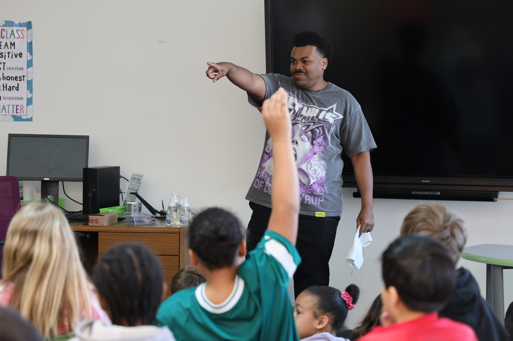 A man stands in front of a group of students and points to one raising their hand